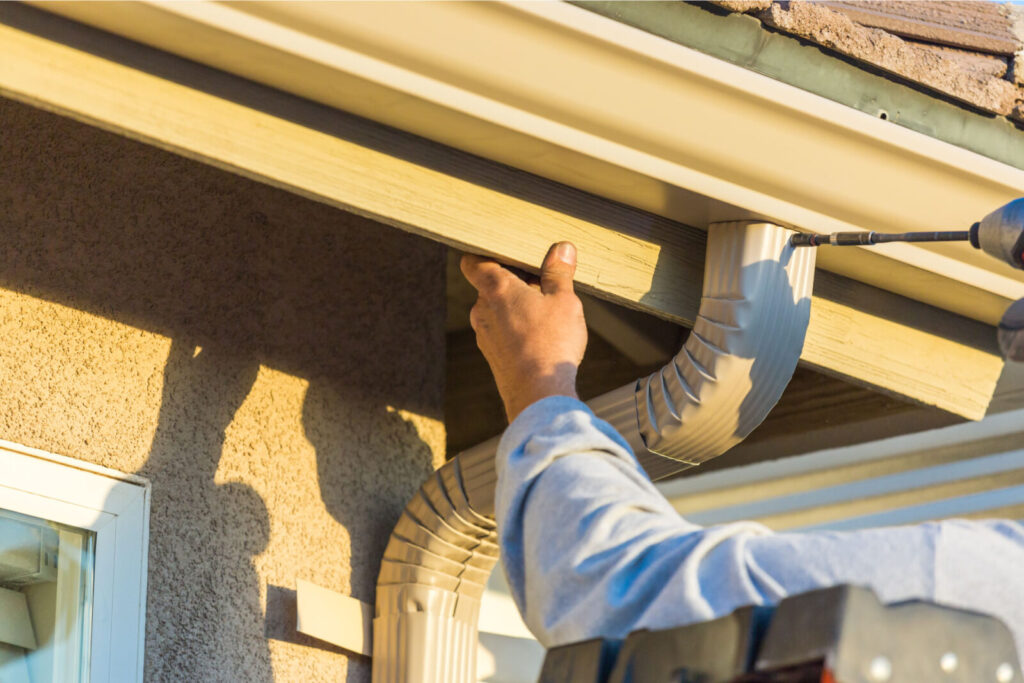 Close-up of a person's hand directing a power washer to clean debris from gutters on a home exterior.